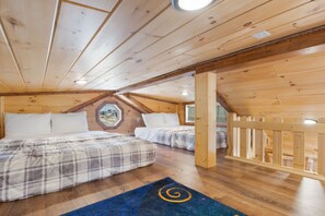 A cabin with a sloped ceiling, wooden walls, two beds with plaid bedding, and a blue rug with a spiral design. - Firelight Cabin | Loft + 2 Baths | Sleeps 8 (Fredericksburg)