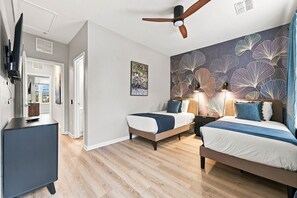 Une chambre avec deux lits, un ventilateur de plafond, un téléviseur fixé au mur et un mur orné d’un motif de feuilles.