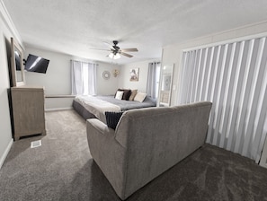 Une chambre à coucher avec un lit, un ventilateur de plafond, une télévision et une fenêtre avec des rideaux.