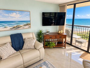 A living room with a beige sofa, a flat-screen TV, a wooden side table, and a view of the ocean through a balcony. - Islandia II Cozy Beachfront 2BR Condo, Ocean & River Views, Hutchinson Island FL (Jensen Beach)