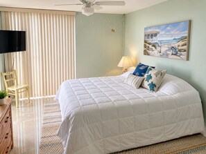 A bedroom with a large bed, a television, a chair, and a painting of a beach scene. - Islandia II Cozy Beachfront 2BR Condo, Ocean & River Views, Hutchinson Island FL (Jensen Beach)