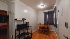 Een compacte kamer met een houten eettafel en stoelen, een zwarte magnetron en een koffiezetapparaat op een plank.