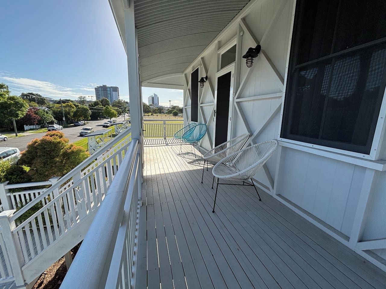 A covered porch with a railing, two chairs, and a view of a parking lot and buildings in the distance.