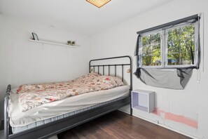 Ein Einzelbett mit floralem Überwurf, ein Fenster mit Blick auf Bäume und ein Regal mit kleinen Pflanzen.