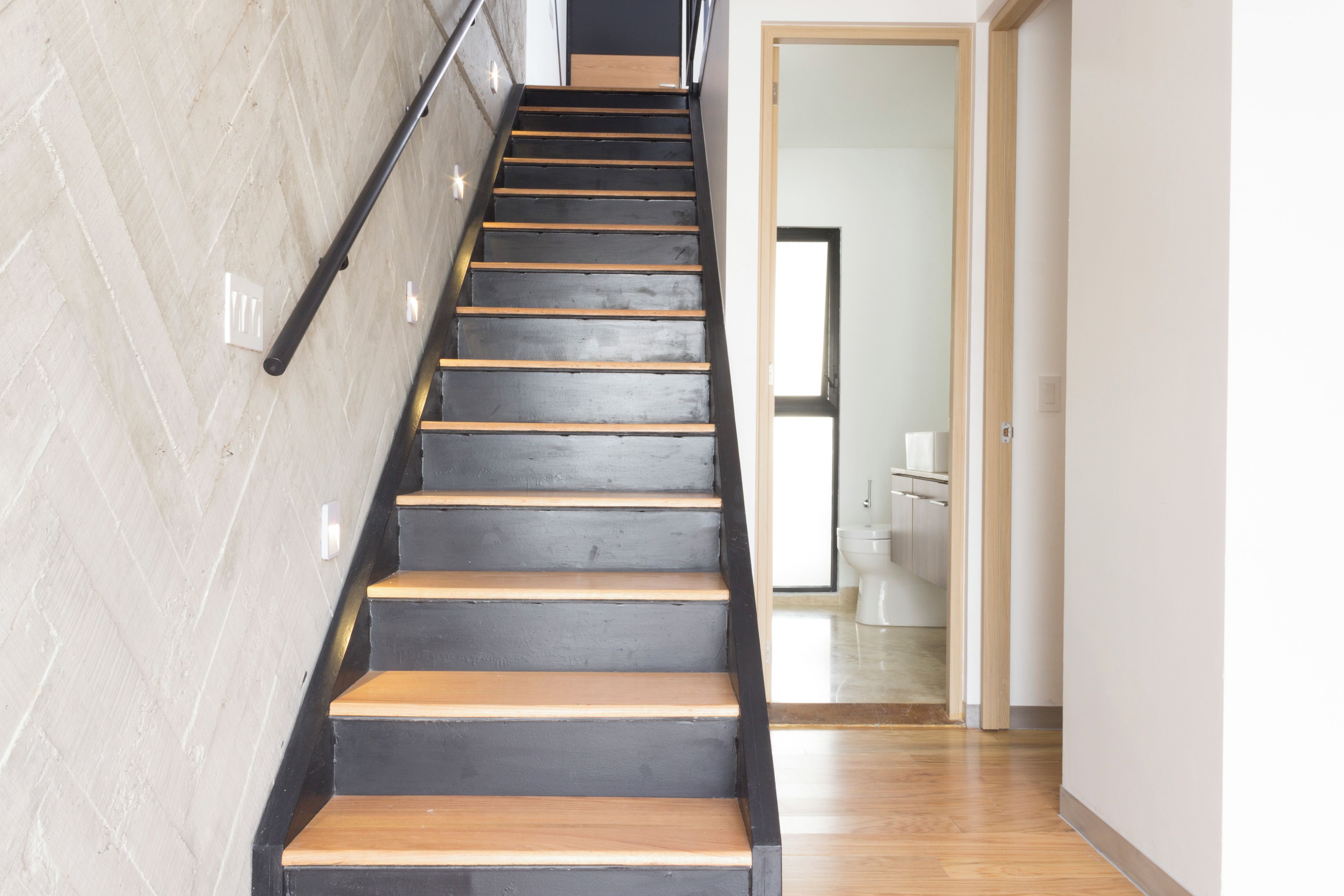 Una escalera de madera con barandillas negras que conduce a un baño con inodoro y lavamanos.