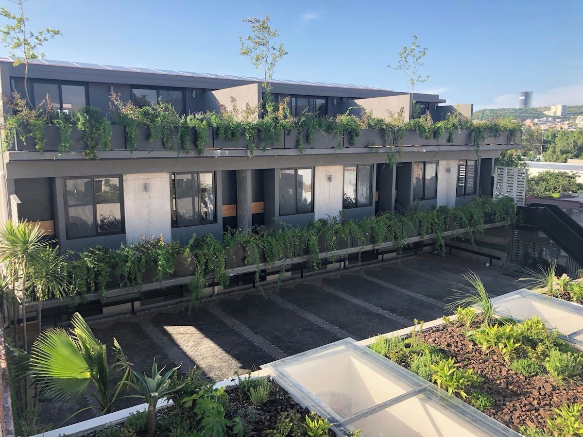 A modern building with multiple floors, large windows, and a rooftop garden with various plants.