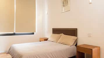 A bedroom with a bed, nightstands, a window with blinds, and a wall-mounted light fixture.