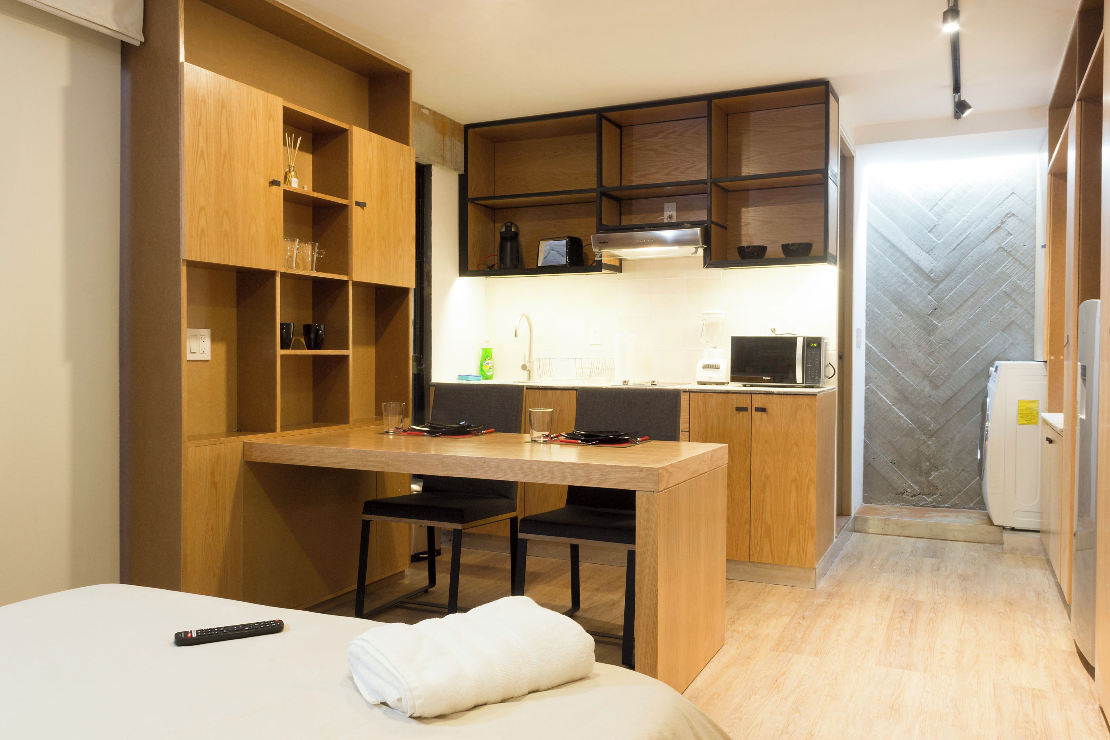 A compact kitchenette with a microwave, coffee maker, and a small refrigerator. There is a wooden dining table with chairs and a bed with a white blanket.