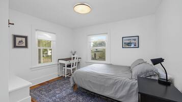 A bedroom with a bed, a desk, two windows, and a nightstand with a lamp.