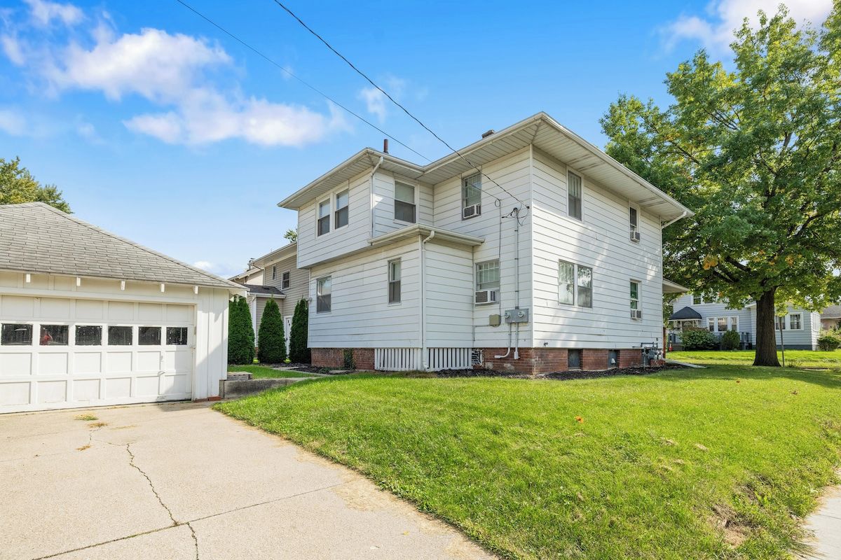 A two-story residential building with a garage, a well-maintained lawn, and a large tree in the front yard.