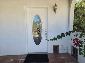 A white door with an oval glass panel, a doorbell, a small vent, and a wall-mounted light fixture.