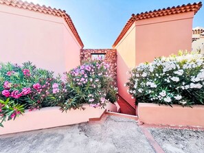 Ein schmaler Gang mit einer Steinmauer und einem kleinen Fenster, umgeben von blühenden, rosa und weißen Pflanzen.
