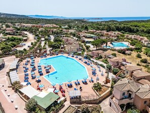 Ein Resort mit einem großen, zentral gelegenen Pool, umgeben von Liegestühlen und Sonnenschirmen, vor dem Hintergrund von Villen im mediterranen Stil und freiem Blick auf das Meer.