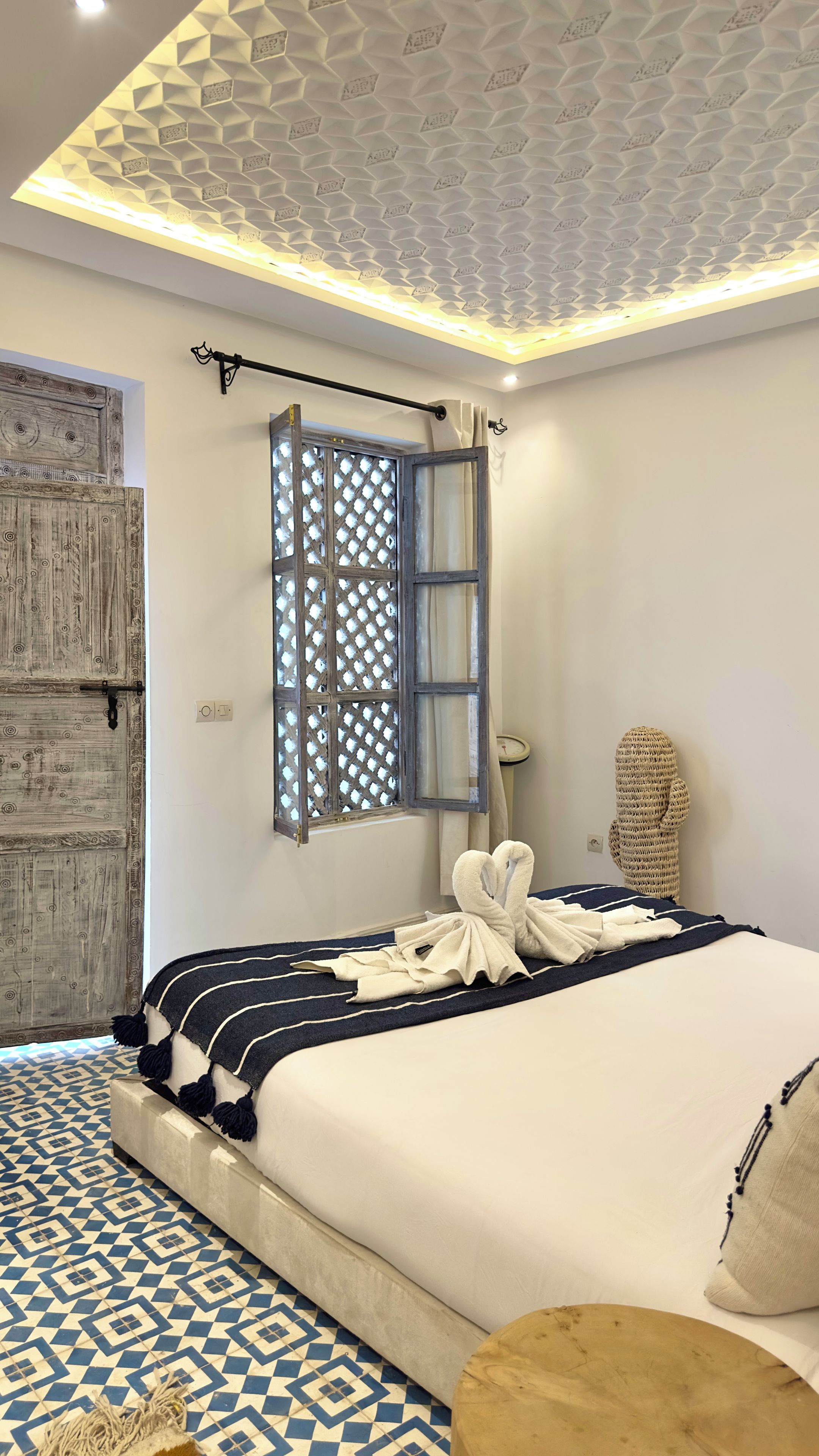 A modern bedroom with a geometric-patterned ceiling, a large bed with a striped blanket, a wooden door, and a window with a lattice design.