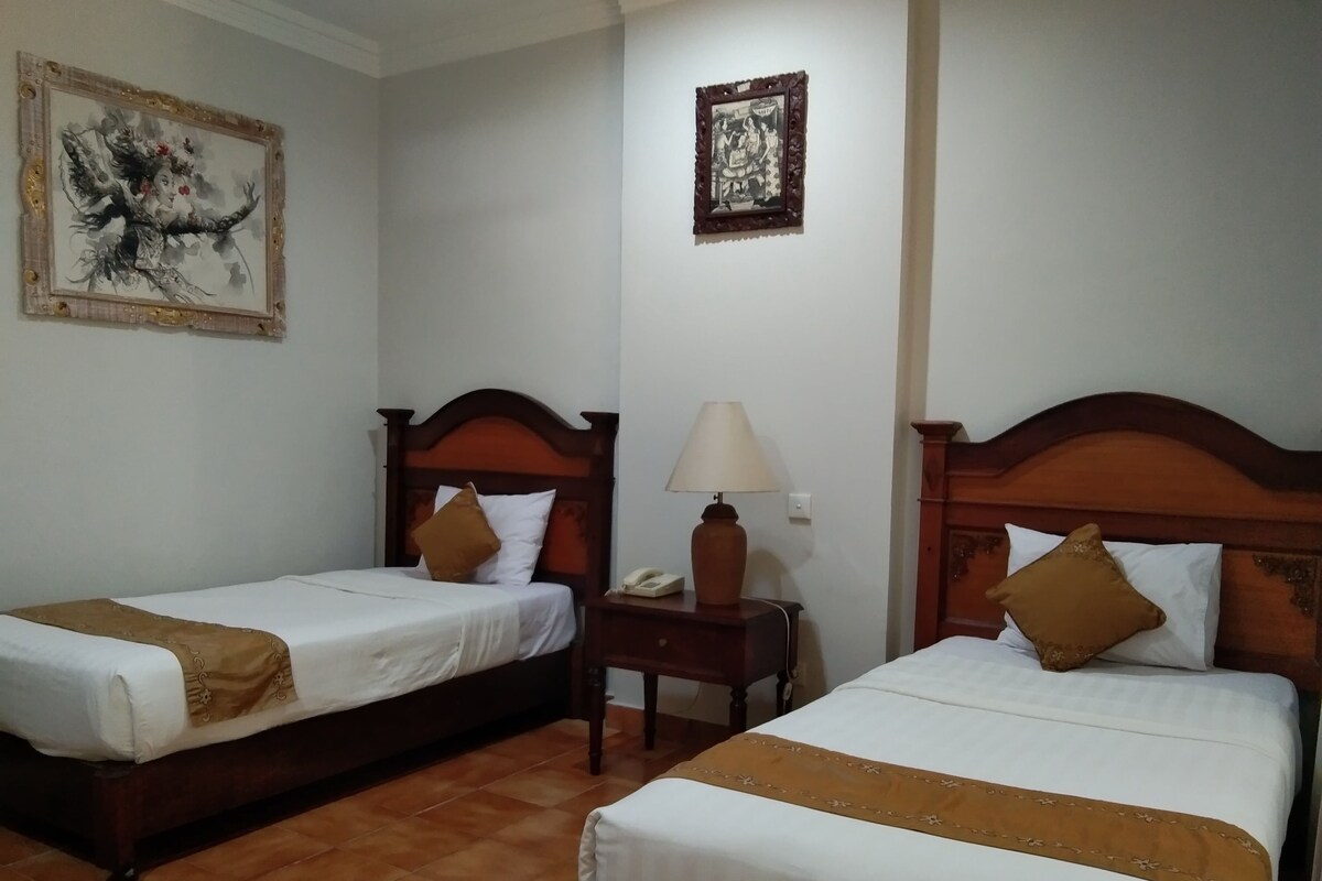 Quarto de hotel com duas camas de solteiro, uma mesinha de cabeceira com abajur e duas obras de arte emolduradas nas paredes.