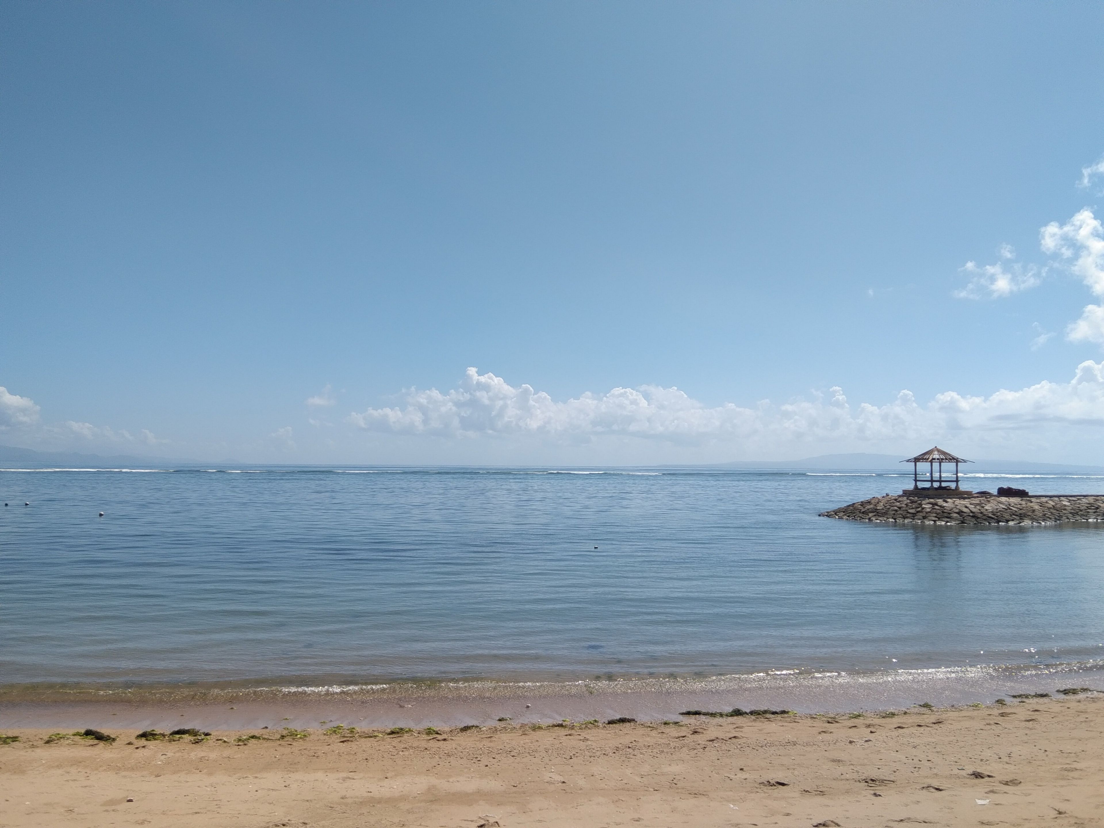 Una playa con aguas tranquilas, un pequeño promontorio rocoso y una estructura tipo mirador.