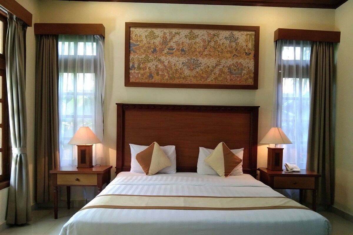 Quarto de hotel com uma cama grande, duas mesas de cabeceira com abajures, um quadro acima da cama e cortinas nas janelas.