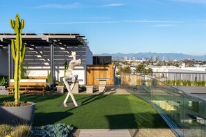 Terraço no telhado com grama sintética, um cacto, um banco de madeira e uma escultura.