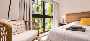Een slaapkamer met een groot raam, een bed met een witte dekbedovertrek en een opgevouwen plaid, een houten hoofdbord, een rieten fauteuil met kussen, en een nachtkastje met een lamp.