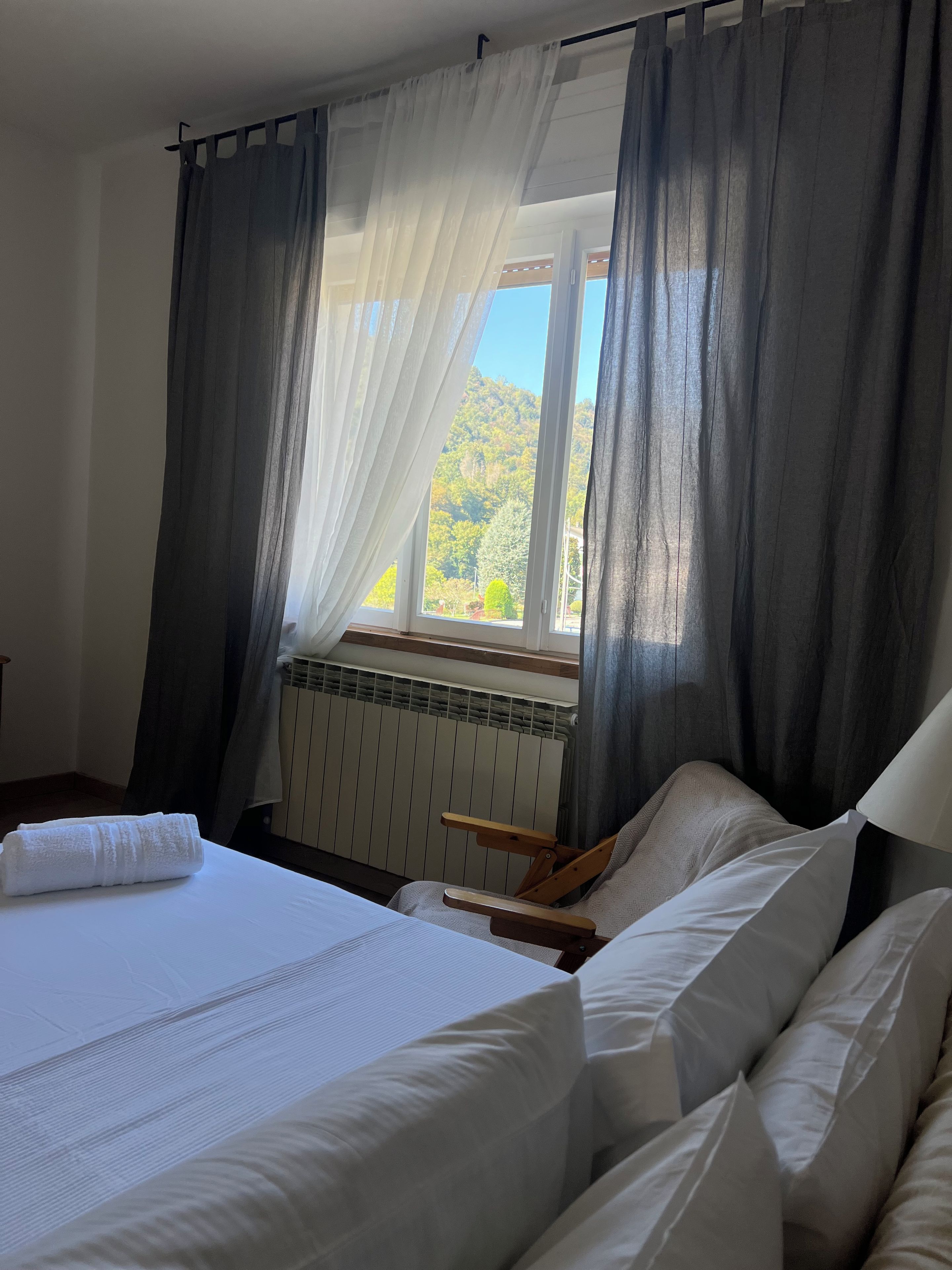 A bedroom with a bed, a window with curtains, a radiator, and a wooden chair.
