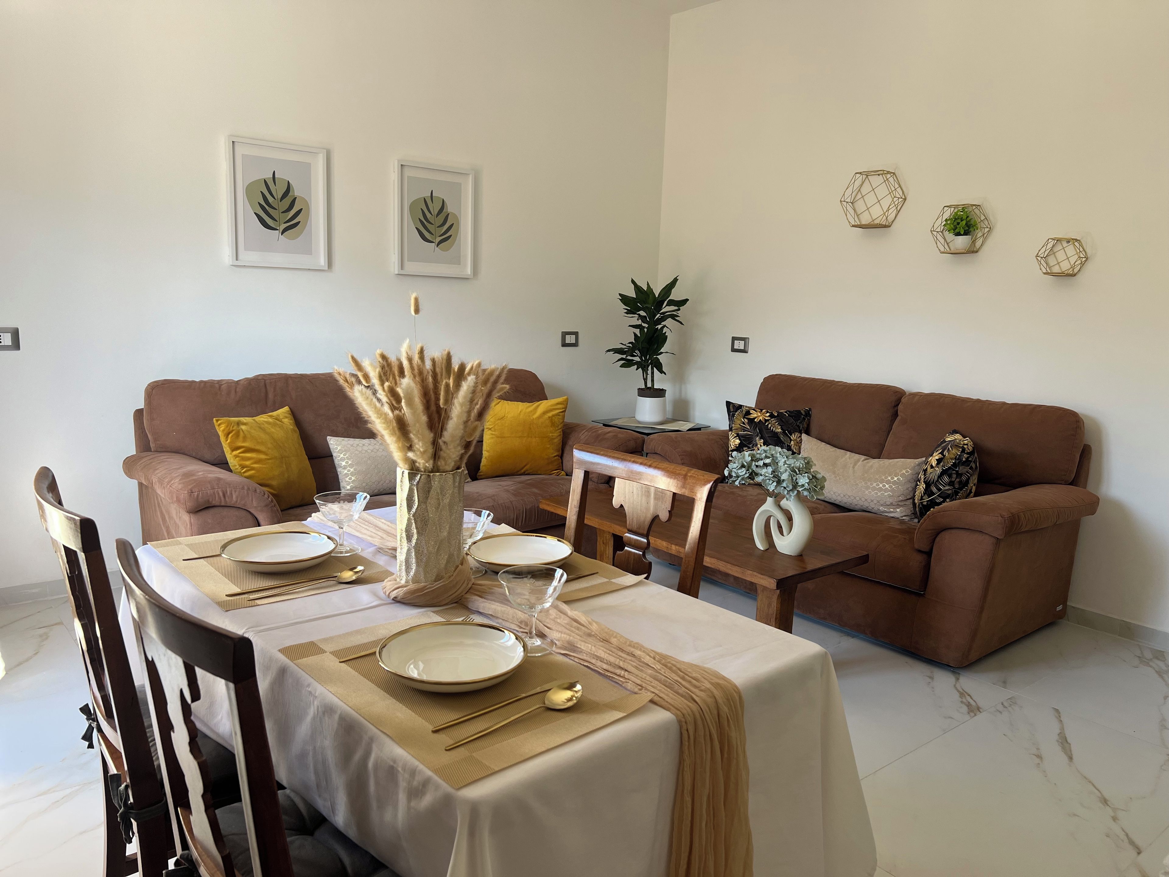 Un coin repas avec une table dressée pour deux personnes, un canapé marron et des œuvres d’art décoratives accrochées au mur.