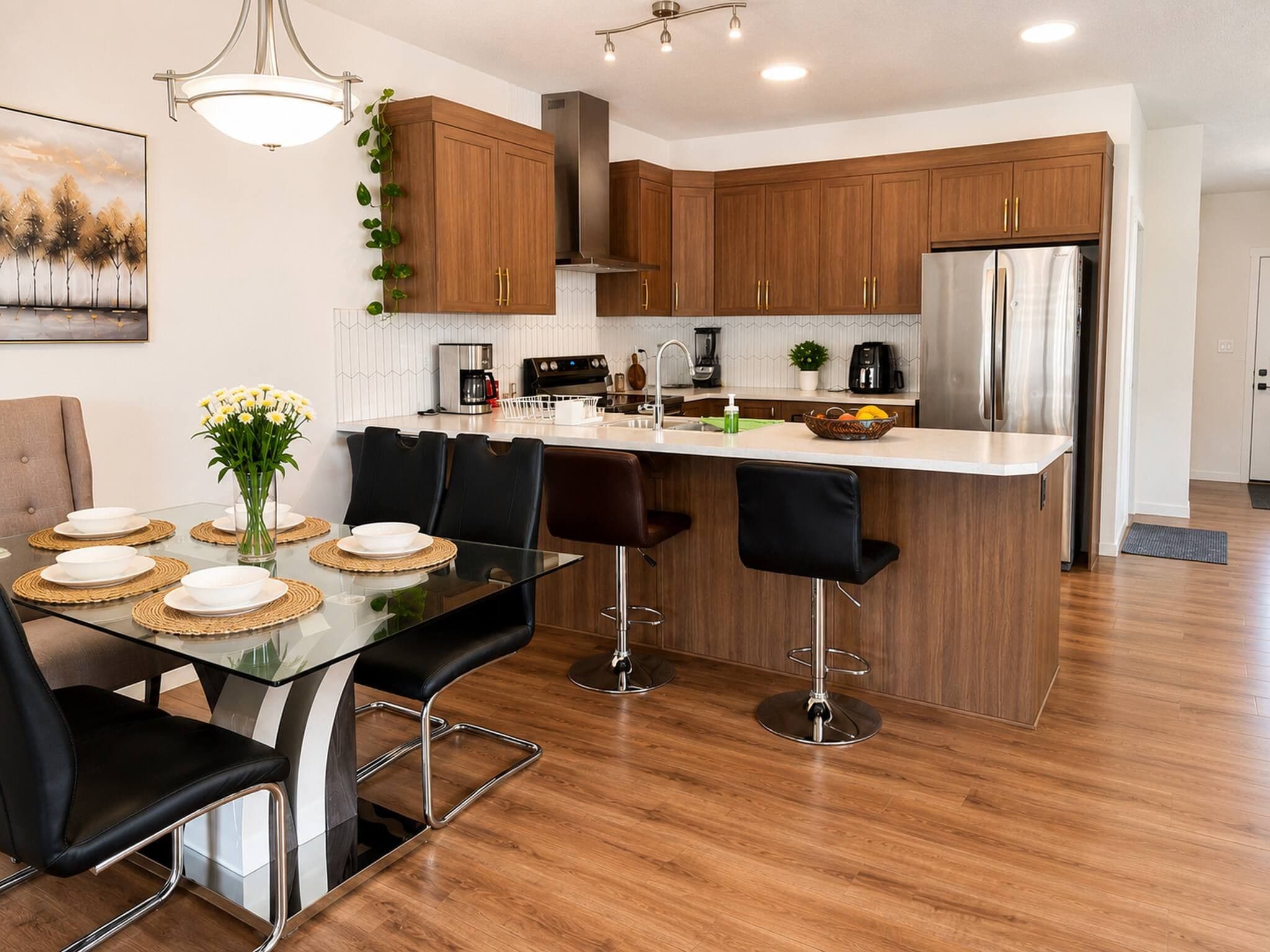 A modern kitchen with wooden cabinets, a stainless steel refrigerator, and a dining area with a glass table and black chairs.