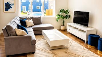 A modern living room with a grey sectional sofa, a white coffee table, a TV on a white stand, a potted plant, and a view of the outside.