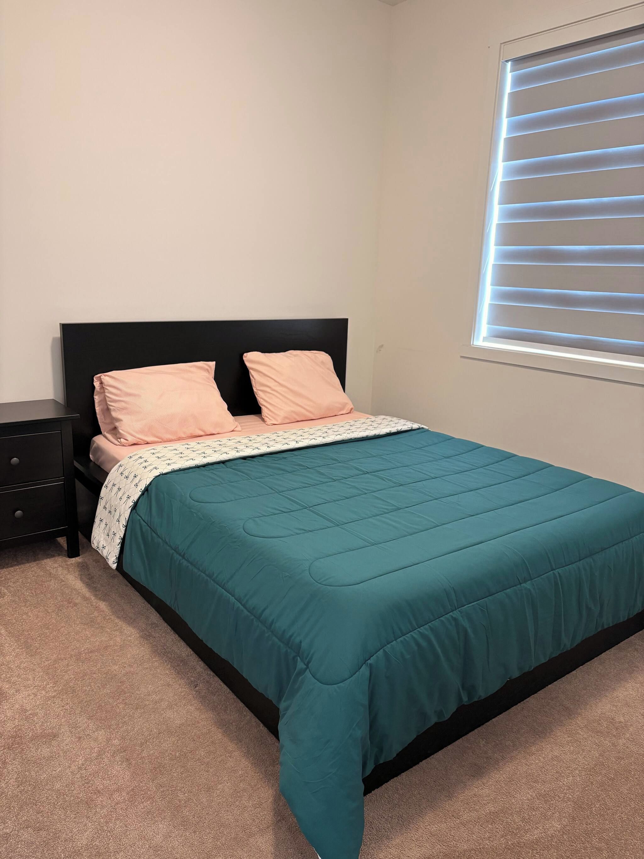 A bedroom with a bed, two pillows, a bedside table, and a window with blinds.