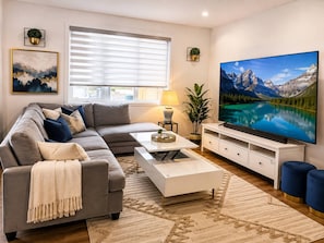 A modern living room with a grey sofa, a coffee table, a TV, and a large window with blinds. - Spaciouscalgaryhome l 4 Bedrooms l Sleeps 8 Plus (Calgary)