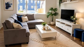 Un soggiorno moderno con un divano angolare grigio, un tavolino da caffè bianco, una TV a schermo piatto su un mobile bianco e un'ampia finestra con persiane.