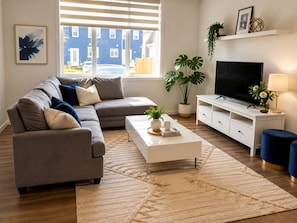 A modern living room with a grey sectional sofa, a white coffee table, a flat-screen TV on a white stand, and a large window with blinds. - Spaciouscalgaryhome l 4 Bedrooms l Sleeps 8 Plus (Calgary)