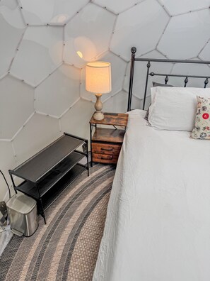 Ein Schlafzimmer mit einem Bett, einem Nachttisch mit Lampe, einem kleinen Holzbeistelltisch, einer Metallbank und einem gemusterten Teppich.