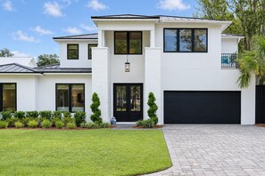 A modern two-story house with a garage, a well-maintained lawn, and a clear sky. - Oak and Palm Cottage – New Listing! Family Retreat Private Pool & Spa | Walk to Village, Parks & Beach (Saint Simons Island)