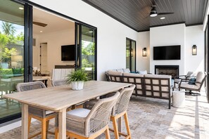 An outdoor living area with a wooden table and wicker chairs, a built-in sofa, and a flat-screen TV mounted on the wall. - Oak and Palm Cottage – New Listing! Family Retreat Private Pool & Spa | Walk to Village, Parks & Beach (Saint Simons Island)