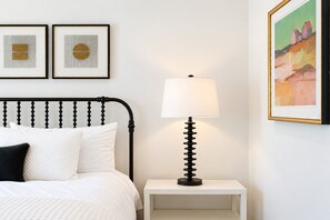 Een keurig opgemaakt bed met witte beddengoed en een zwart hoofdeinde, een nachtkastje met een lamp, en twee ingelijste kunstwerken aan de muur.