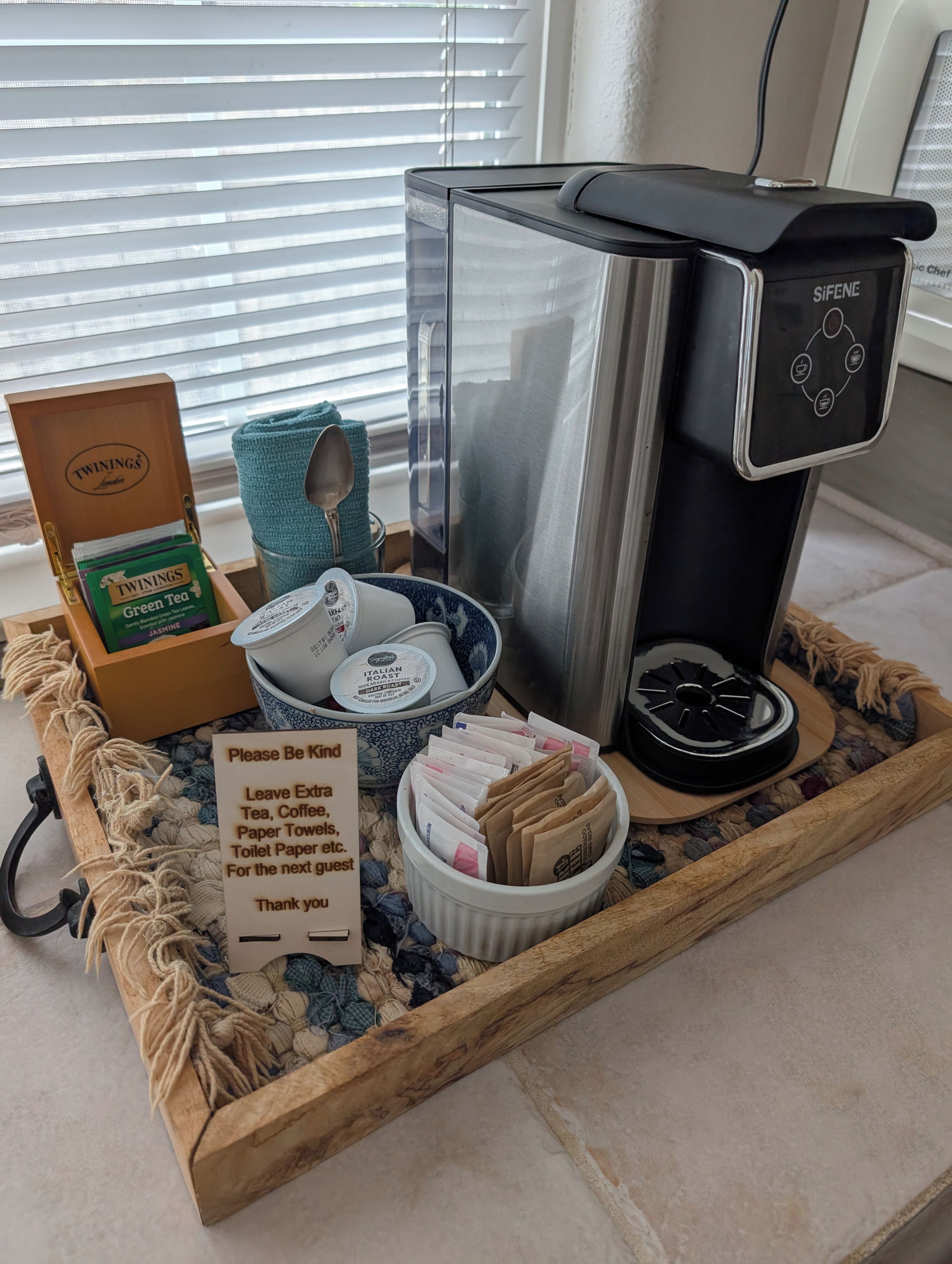 Angolo caffè con macchina da caffè, tè Twinings, un vassoio con tè in busta e un cartello che invita gli ospiti a lasciare tè e caffè in eccesso. Un vassoio contiene anche un cucchiaino e una tazza.