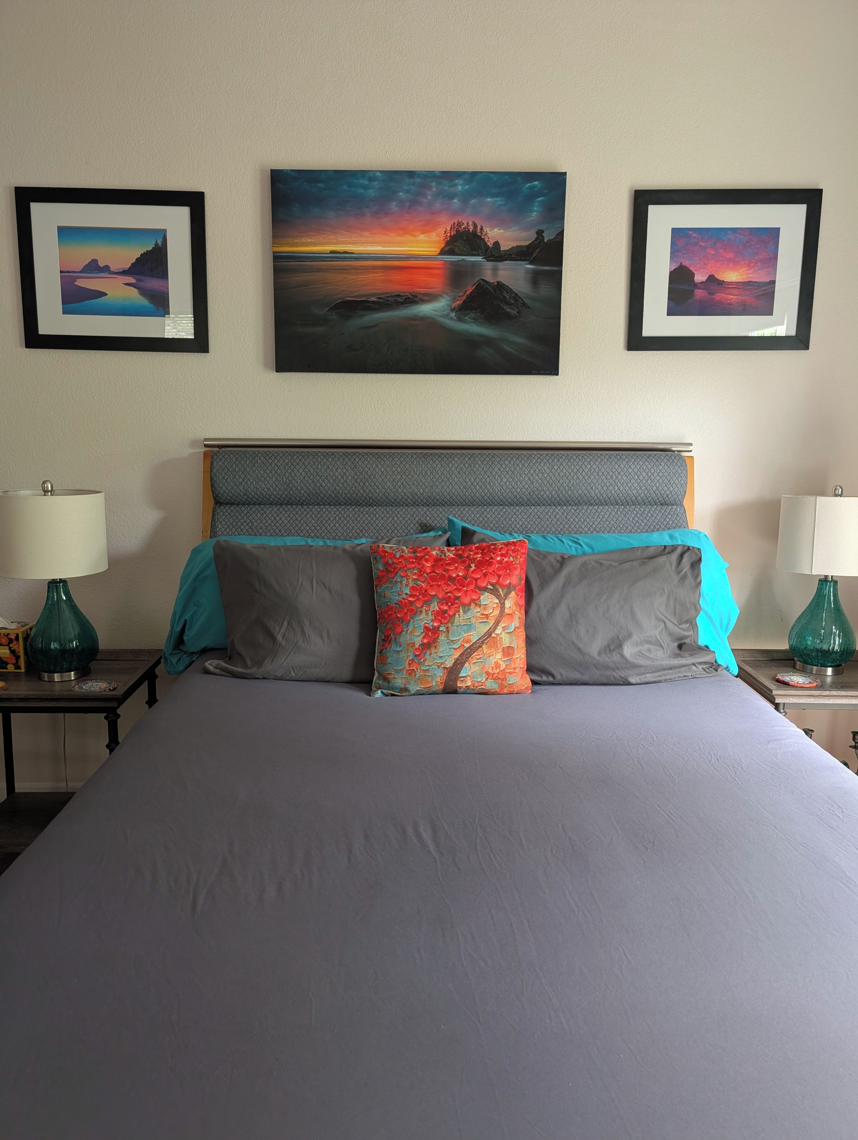 A bed with a grey mattress, two pillows, and a headboard. There are two nightstands with lamps and a framed picture above the bed.