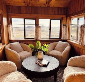 A cozy room with wooden walls and large windows, featuring a round table with a plant and books. - Cozy Log Cabin at Blue Skies Ranch, minutes to Palmer Lake (Larkspur)