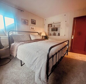 Un dormitorio con estructura de cama metálica, una mesita de noche, una lámpara, un estante con objetos decorativos y una puerta.