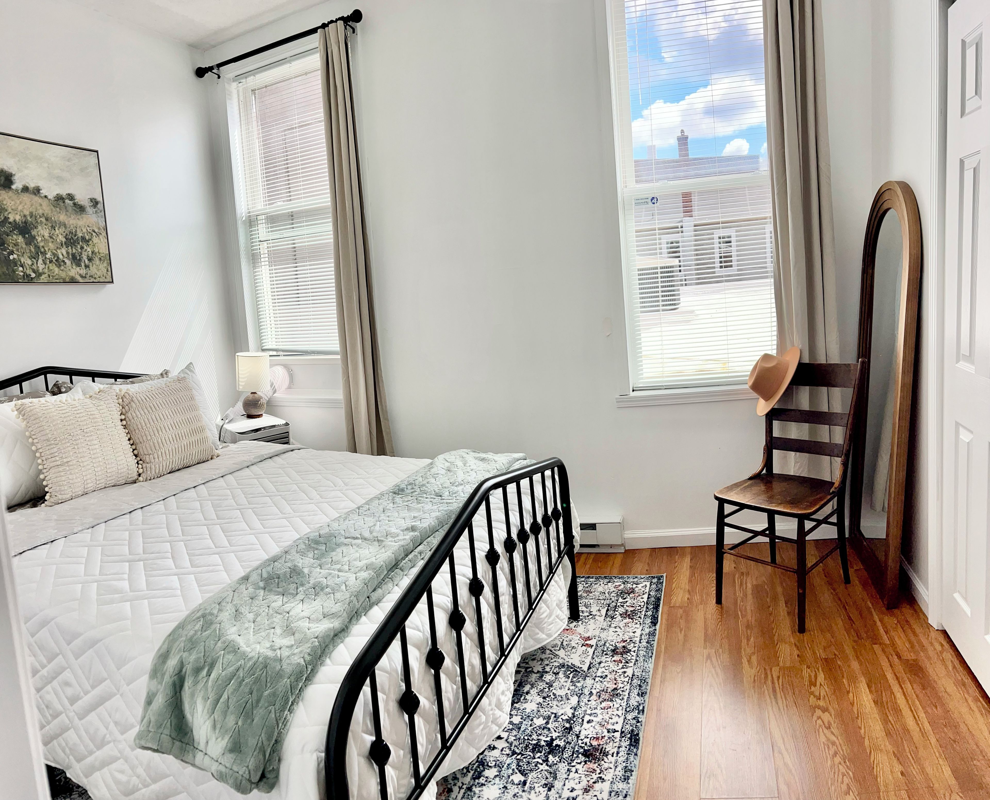 A bedroom with a bed, a chair, a window with blinds, a painting on the wall, and a rug on the floor.