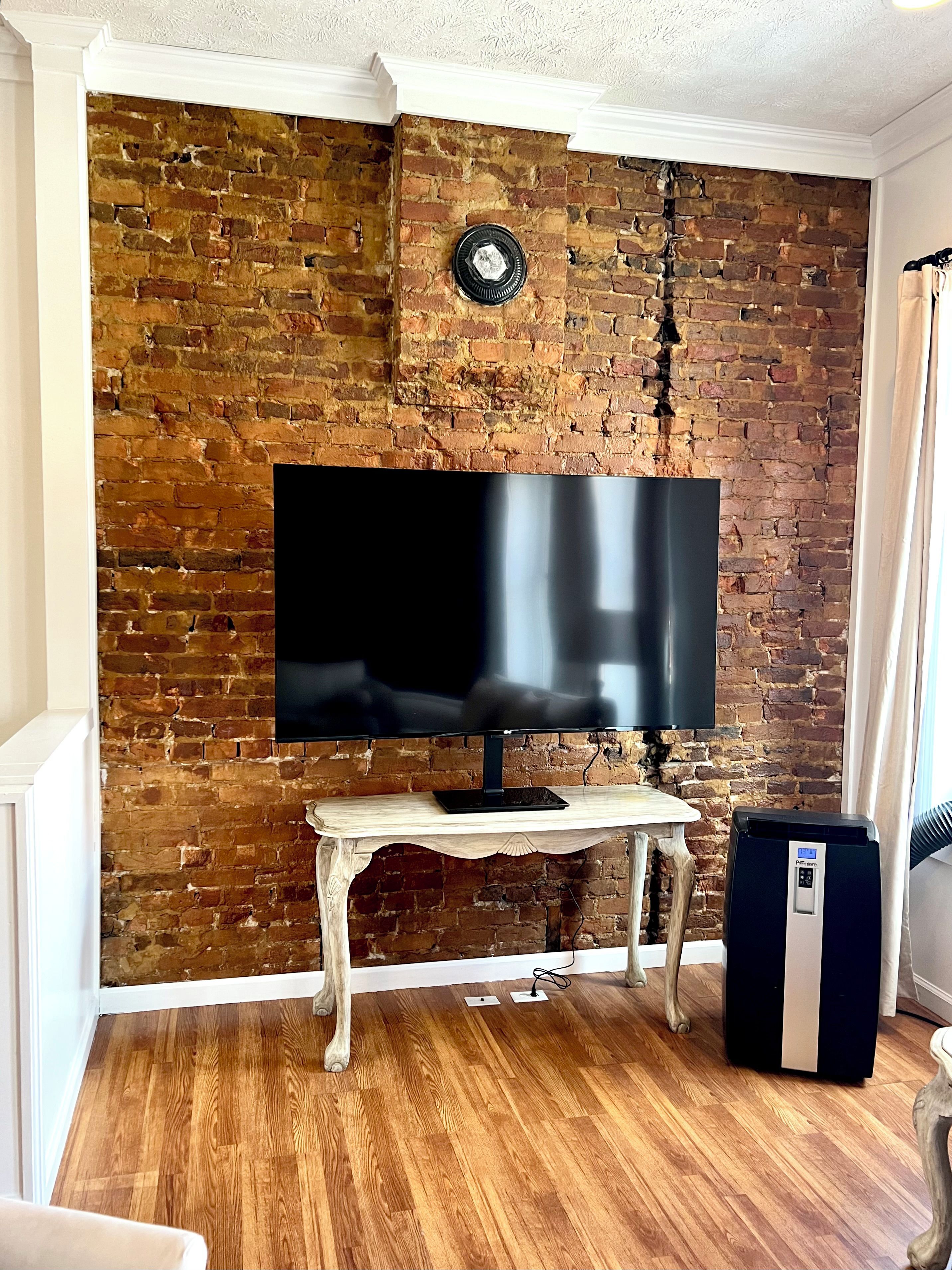 A room with a brick wall, a flat-screen TV on a wooden stand, and a black portable heater.