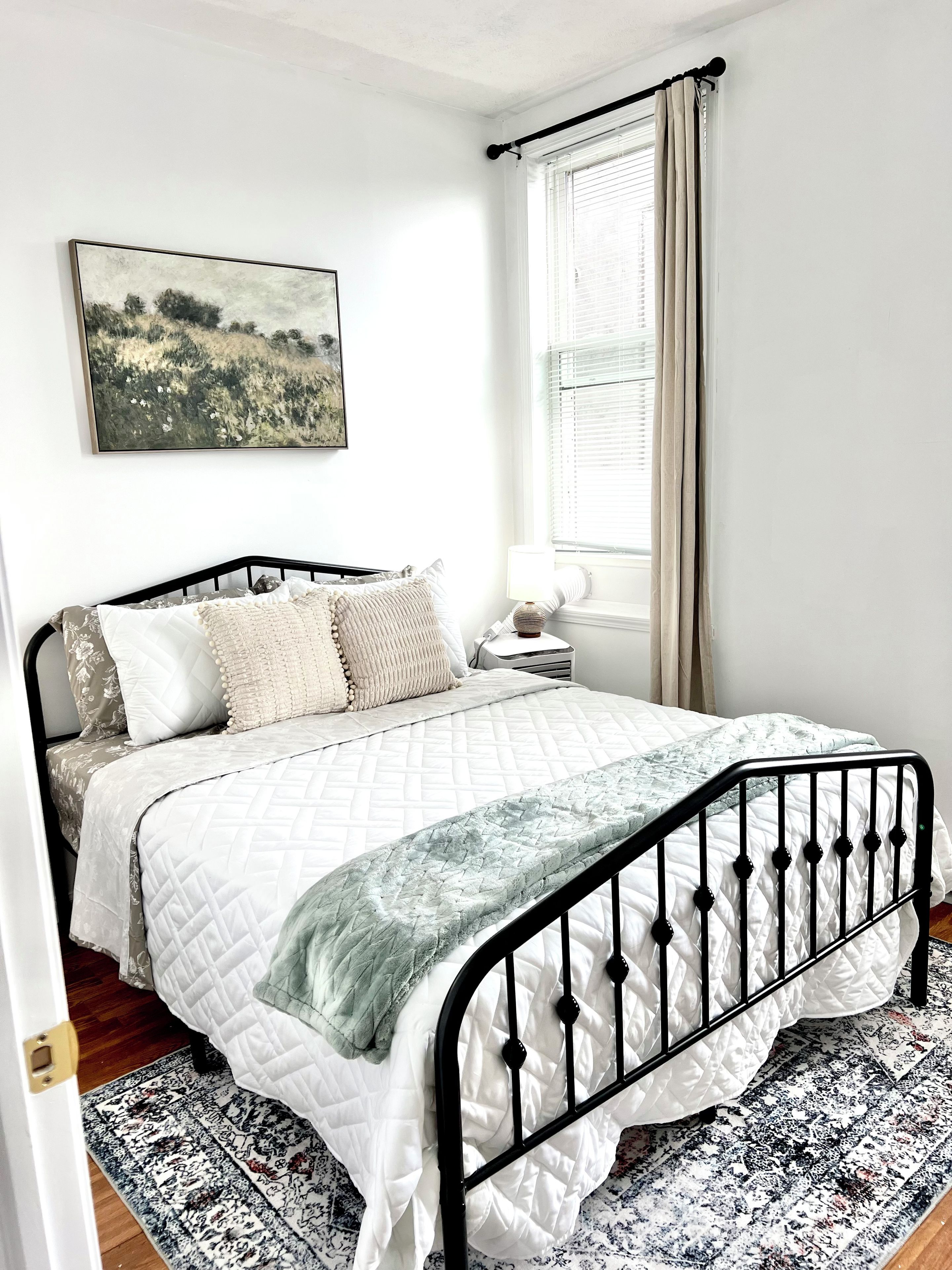 A bedroom with a bed, a bedside table, a lamp, a window with curtains, and a painting on the wall.