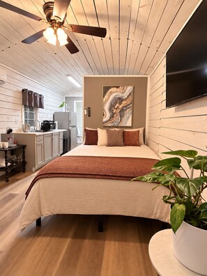 A bedroom with a bed, a ceiling fan, a painting, and a TV on the wall.