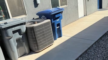 A building exterior with a blue trash bin, a grey dumpster, and a window with a screen.