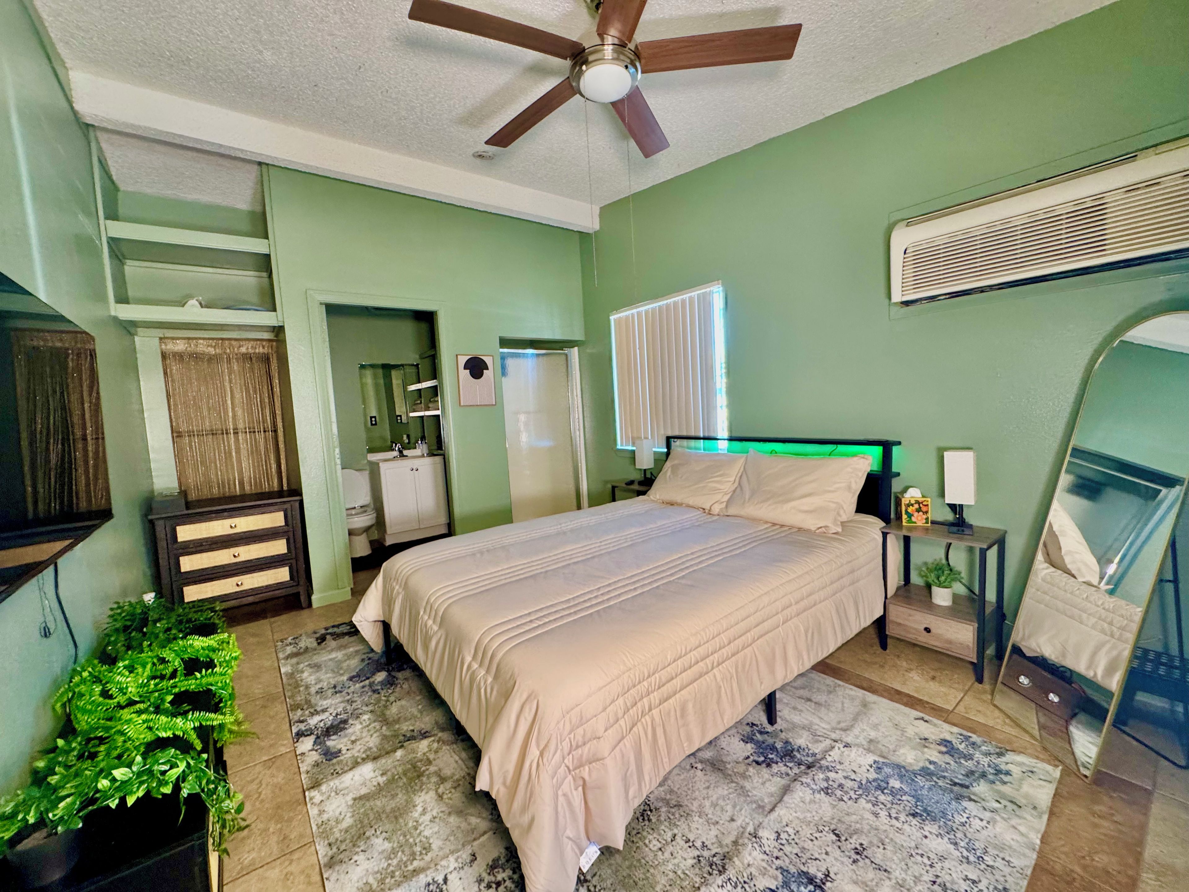 A bedroom with a bed, a ceiling fan, a bedside table with a lamp, a mirror, and a window with blinds.