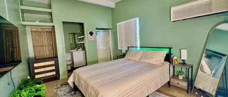 A bedroom with a bed, a ceiling fan, a bedside table with a lamp, a mirror, and a window with blinds.