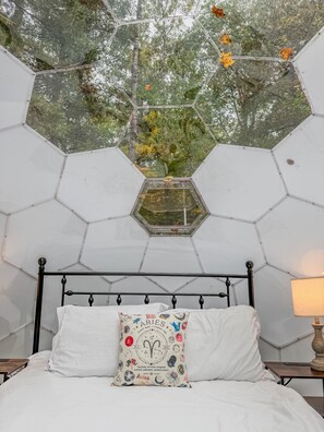 Un plafond hexagonal agrémenté d’une lucarne, un lit avec un matelas blanc et un oreiller orné de motifs du zodiaque, ainsi qu’une table de chevet avec une lampe.