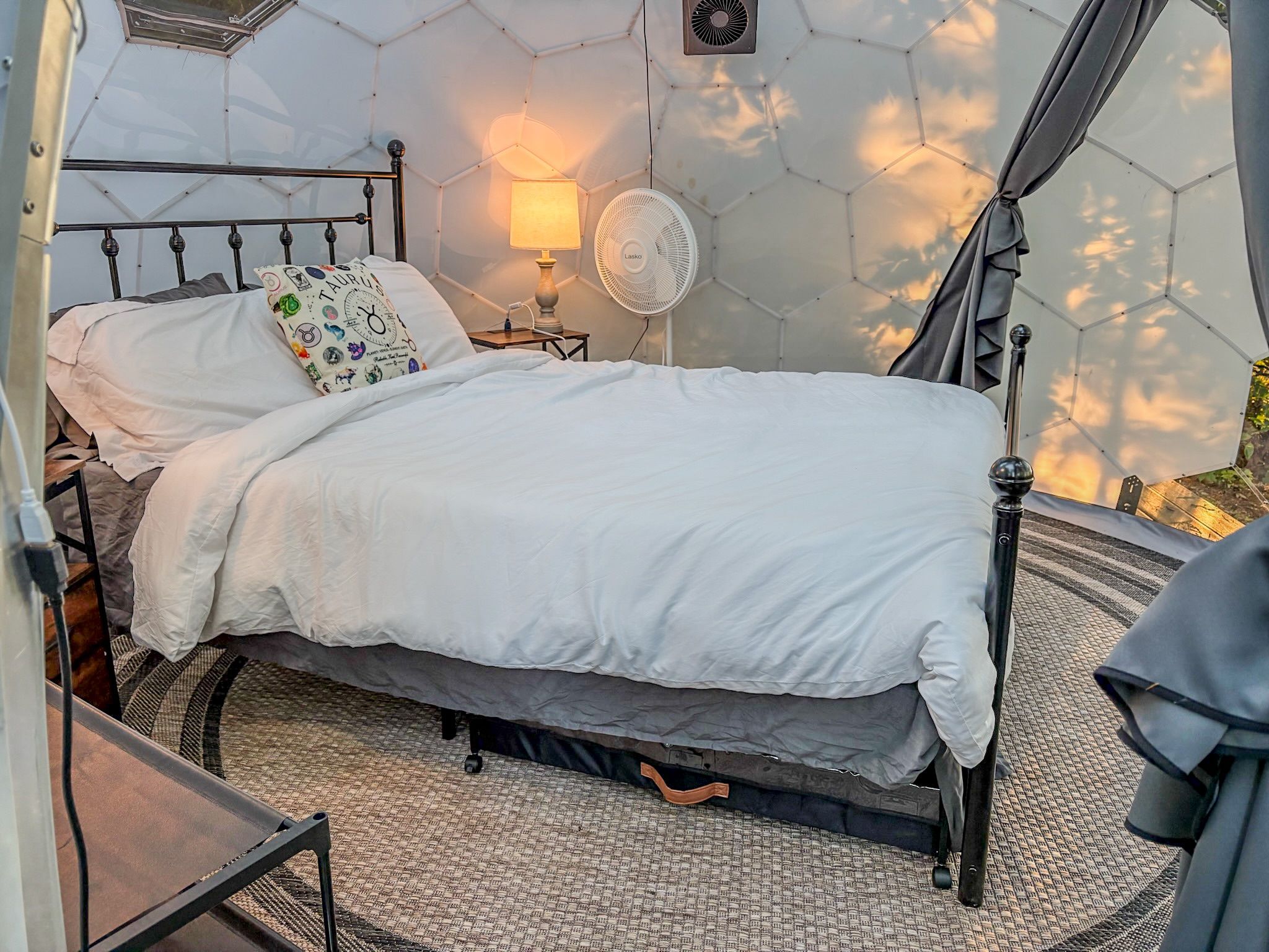 Ein Bett mit weißer Bettdecke, ein Nachttisch mit Lampe und ein an der Wand befestigter Ventilator.