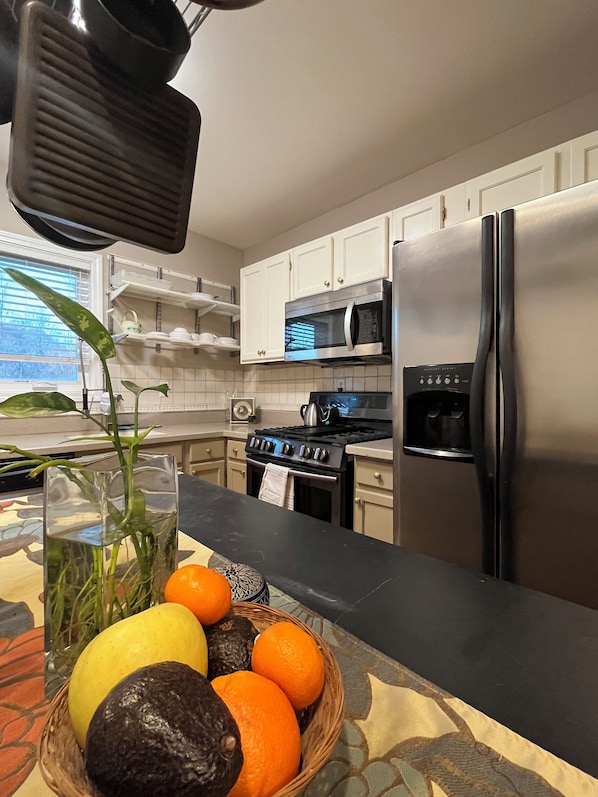 A kitchen with white cabinets, a stainless steel refrigerator, a microwave, and a stove. There is a fruit bowl on the counter with oranges and a lemon.