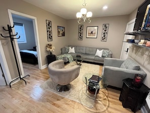 A living room with a grey sofa, a glass coffee table, a grey armchair, a black side table with a lamp, and a chandelier. - 2-bedroom apartment in beautiful Northbrook with AC and comfort (Northbrook)
